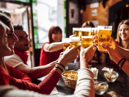 A group of friends is sitting indoors, raising glasses of beer in a toast, creating a lively and cheerful atmosphere.