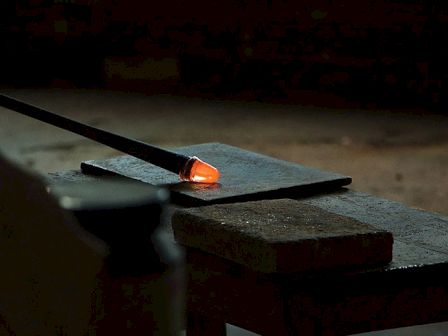 The image shows a glassblowing rod with red-hot molten glass at the tip resting on a flat surface in a dimly lit environment.