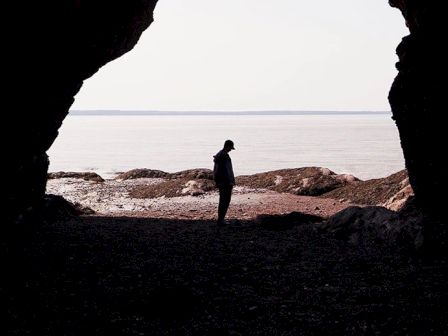 A silhouetted person stands at the entrance of a cave, overlooking a calm, rocky shoreline with water in the distance.