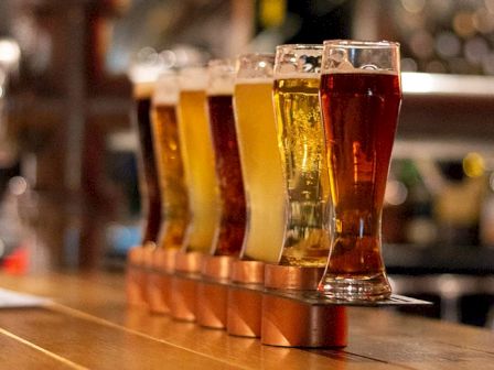 The image shows a row of beer glasses filled with various types of beer, each a different color, resting on a wooden counter in a bar setting.