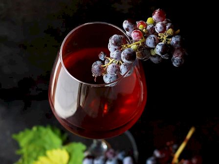 A glass of red wine with a bunch of grapes resting on the rim, set against a dark background with green leaves.