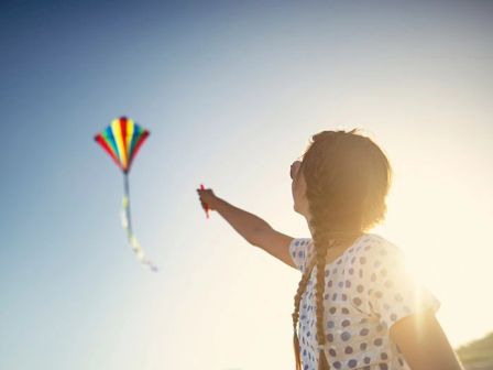 A person holding a string attached to a colorful kite flying in the sky. The sun is shining brightly behind them, creating a radiant atmosphere.