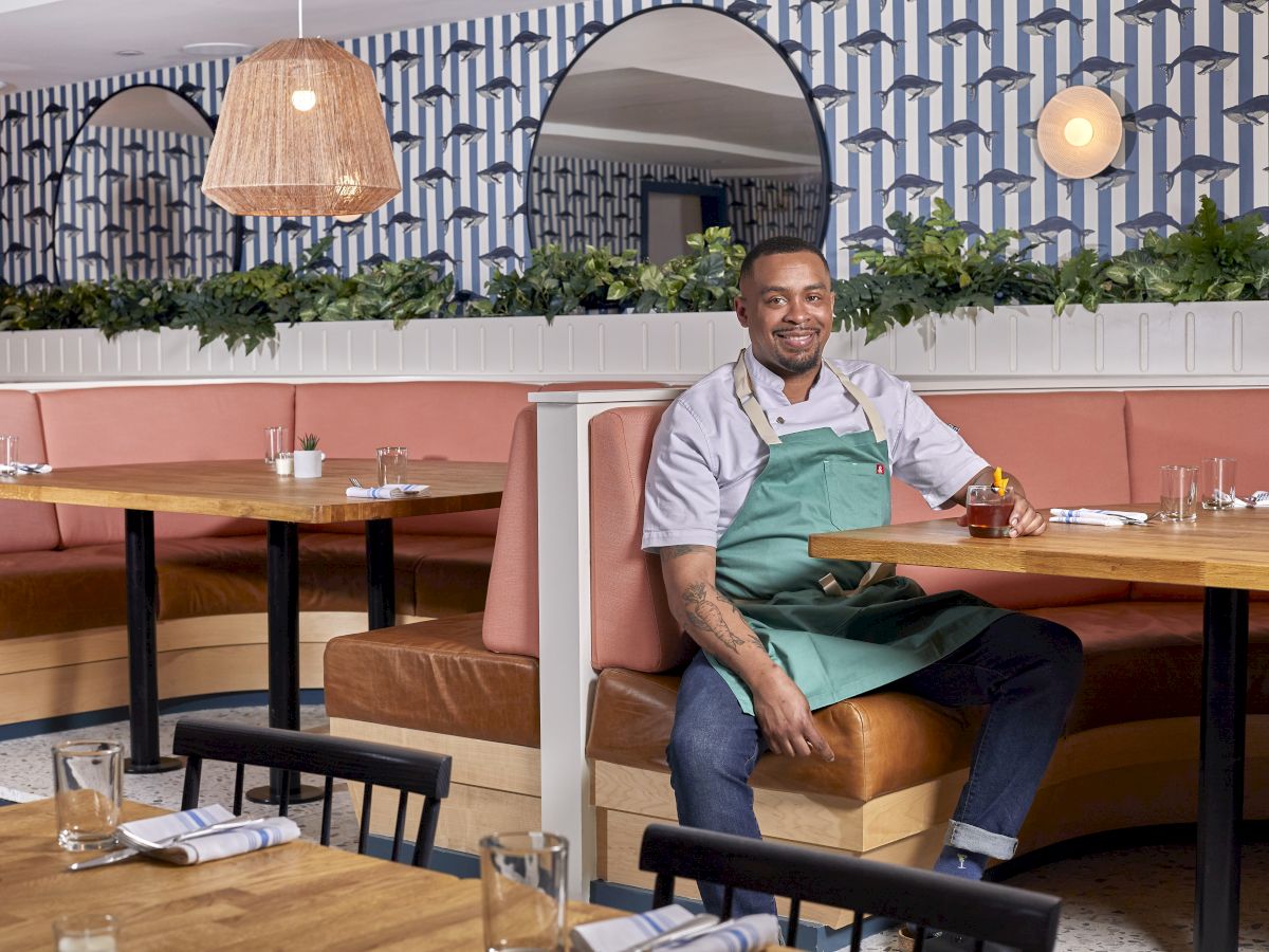 A smiling man in a green apron sits at a booth in a stylish restaurant with pink seating, wooden tables, plants, and pendant lights.