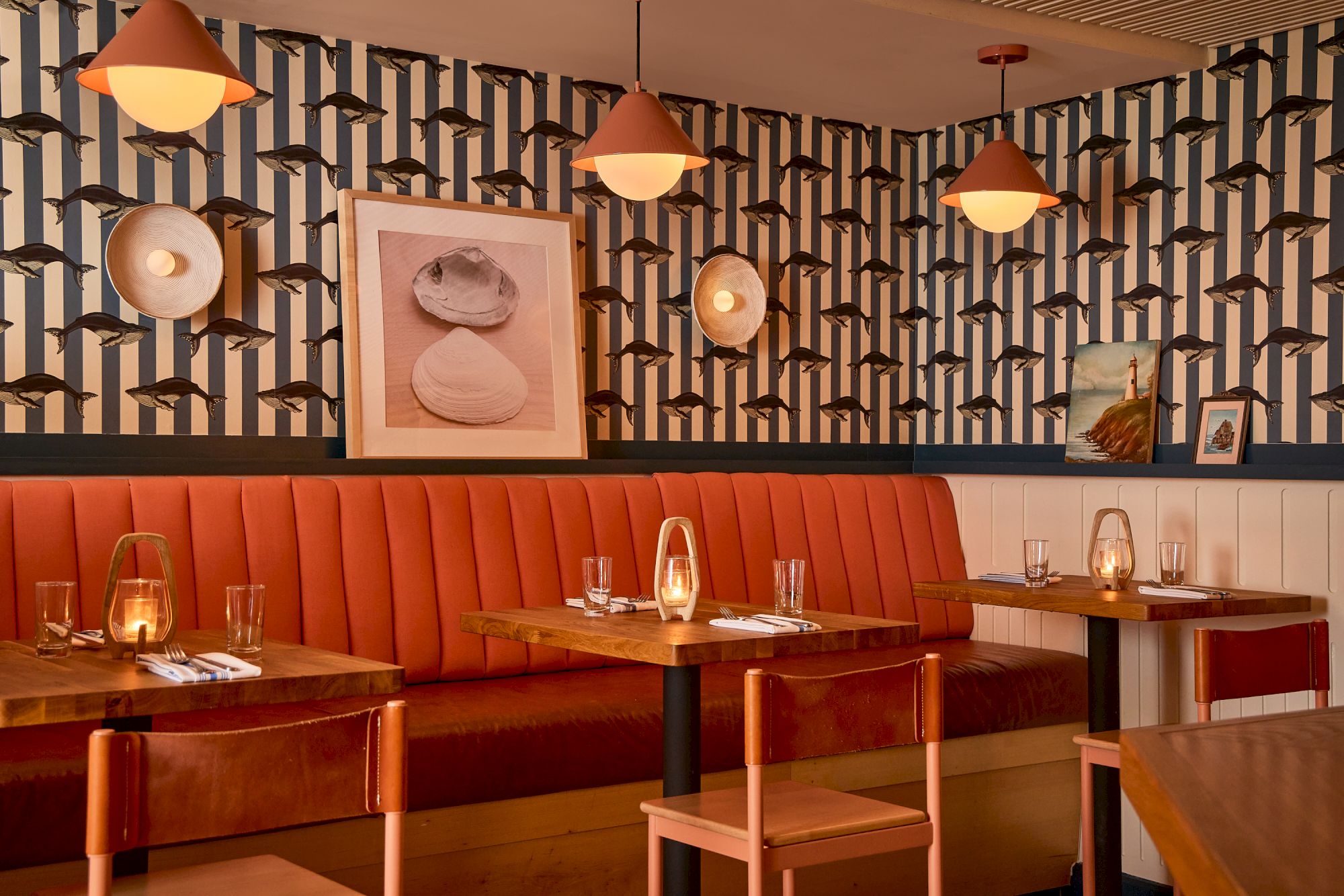 A stylish dining area with a red-orange booth, wooden tables, pendant lights, patterned wallpaper, framed artwork, and cozy table settings.