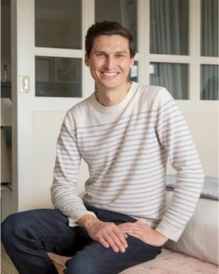 A man wearing a striped sweater and dark pants sits and smiles indoors, with glass-paneled doors in the background.