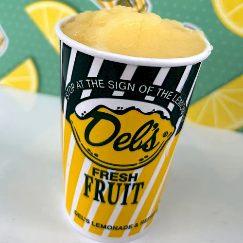 A striped lemon cup with yellow fruit slush, bold &ldquo;Deli&rsquo;s&rdquo; logo, and fresh fruit graphics in the background, topped with a foamy scoop.