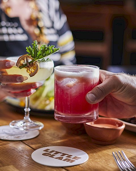 A person hands a pink cocktail to another across a wooden table, with savory dishes and lime garnish in the background.