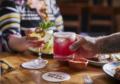 A person hands a pink cocktail to another across a wooden table, with savory dishes and lime garnish in the background.
