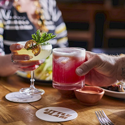 A person hands a pink cocktail to another across a wooden table, with savory dishes and lime garnish in the background.