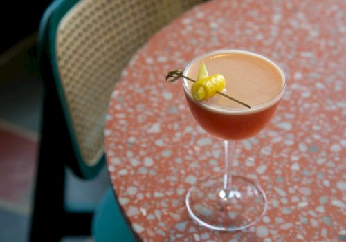 A cocktail with a cherry/lemon garnish in a coupe glass sits on a pink terrazzo table; a chair with a woven back is in the background.
