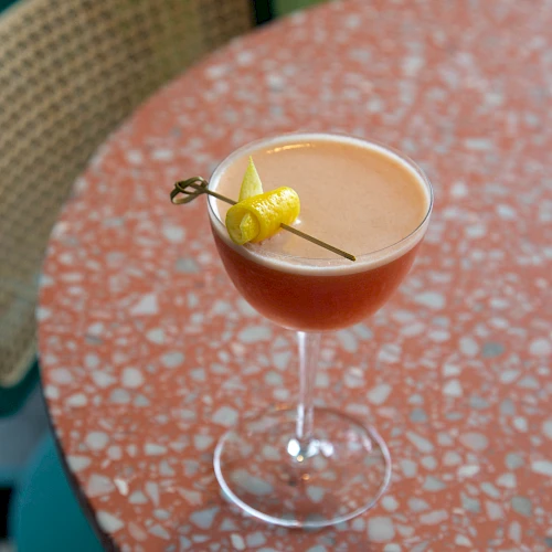 A cocktail with a cherry/lemon garnish in a coupe glass sits on a pink terrazzo table; a chair with a woven back is in the background.