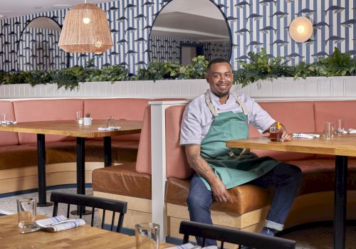 A smiling man in a green apron sits at a booth in a stylish restaurant with pink seating, plants, warm lighting, and patterned wallpaper.