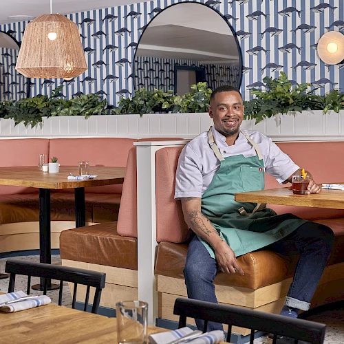A smiling man in a green apron sits at a booth in a stylish restaurant with pink seating, plants, warm lighting, and patterned wallpaper.