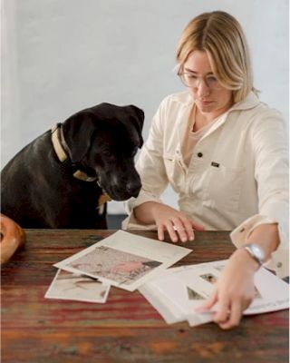 A person is sitting at a table with a black dog, reviewing various papers or photographs laid out in front of them, wearing glasses and white attire.