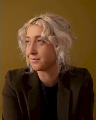 A person with blonde wavy hair in a black top and dark blazer poses against a yellow background, looking to the left with a relaxed expression.