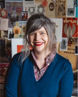 A person with long hair and bright lipstick smiles in a room full of posters and pictures pinned on the wall behind them. The person is wearing a blue top.