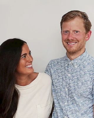 A woman and a man are standing close together, smiling at each other in front of a plain background.