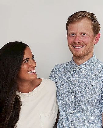 A woman and a man are standing close together, smiling at each other in front of a plain background.