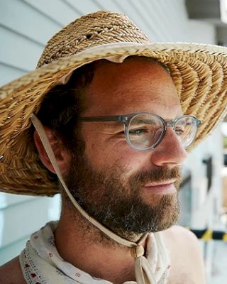 A bearded man wearing glasses is seen in a straw hat with a chin strap tied under his chin, standing outside near a light blue wall.