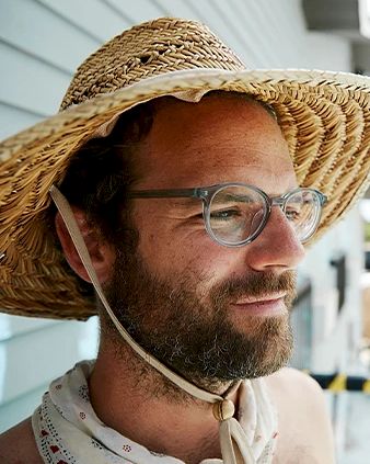 A bearded man wearing glasses is seen in a straw hat with a chin strap tied under his chin, standing outside near a light blue wall.