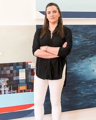 A person stands with arms crossed, wearing a black shirt and white pants in front of a painting that appears to depict a nautical theme.