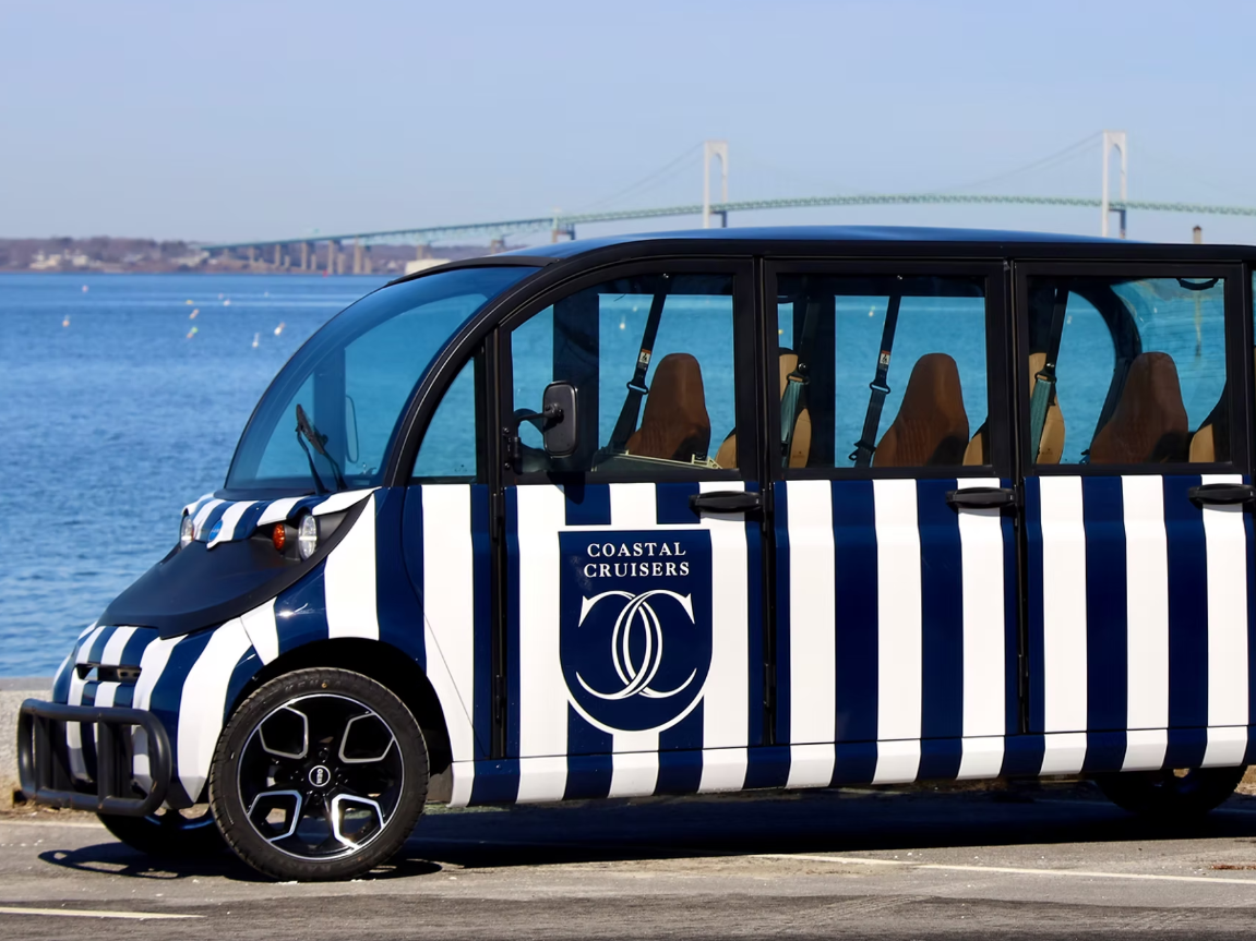 A striped blue-and-white passenger shuttle with open side windows parked by the water, &ldquo;Coastal Cruisers&rdquo; logo on the door.
