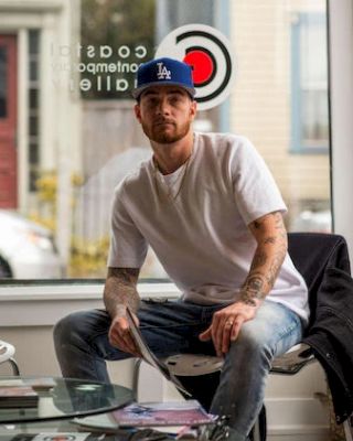 A man in a white t-shirt and blue cap sits at a glass table, holding a magazine, near a window with a target design.