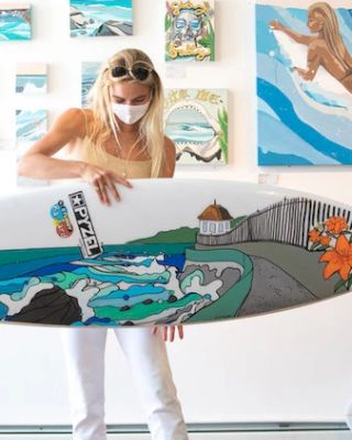 A person wearing a mask and sunglasses holds a decorated surfboard in an art gallery with colorful paintings in the background.