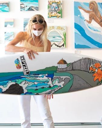 A person wearing a mask and sunglasses holds a decorated surfboard in an art gallery with colorful paintings in the background.