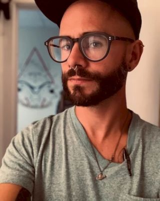 A bearded person with glasses and a cap takes a selfie, wearing a light green shirt and standing indoors with blurred background decor.