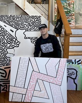 The image shows a person in a cap and black shirt standing beside abstract art pieces in a room, with stairs and more art in the background.