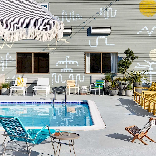 A gray house with string lights, a small pool, outdoor chairs, umbrellas, and quirky white doodles across the wall under a clear blue sky.