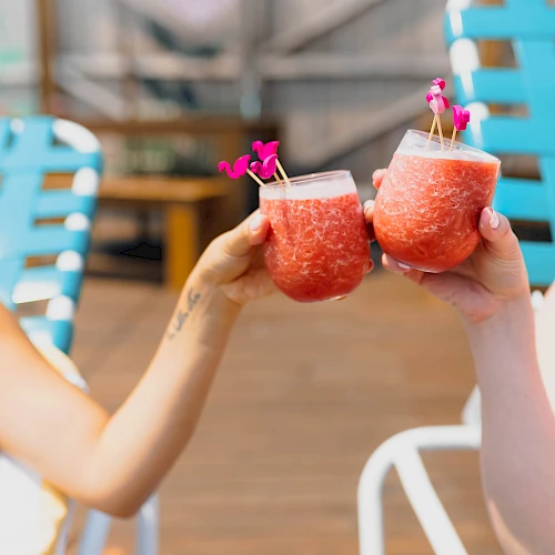 Two people clinking pink grapefruit cocktails with tiny umbrellas, cheerful poolside vibe, blue chairs in the background.