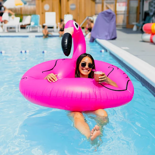 A woman floats in a bright pink flamingo pool float at a sunny hotel pool, wearing sunglasses and enjoying the water.
