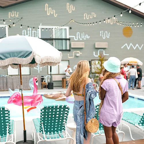 Two women stand by a poolside with pastel decor; lounge chairs, a flamingo float, string lights, and a teal-green building backdrop.