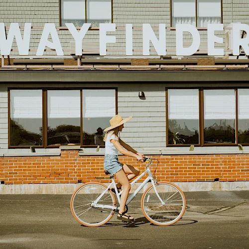 A person riding a bicycle past a building with a large &ldquo;WAYFINDER&rdquo; sign on top, sunny day.