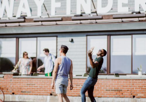People standing outside a brick building with large windows; someone practices a balance pose on a wooden board while others chat nearby.