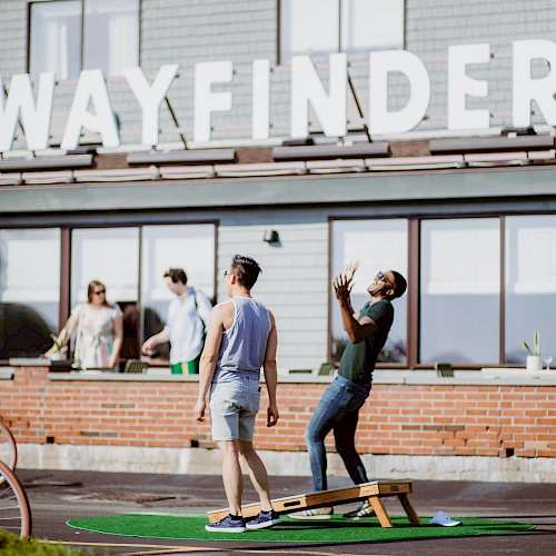 People standing outside a brick building with large windows; someone practices a balance pose on a wooden board while others chat nearby.