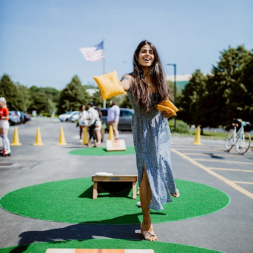 A cheerful girl throws a yellow glove in a parking lot mini-golf area, balancing on a green mat with wood blocks and cones around her.