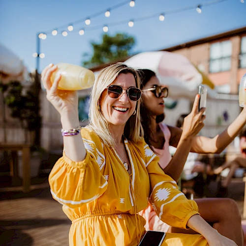 Two friends at an outdoor party sip drinks, one holds a roll, smiling in a yellow dress, sunny vibes, string lights above, phones in hand.