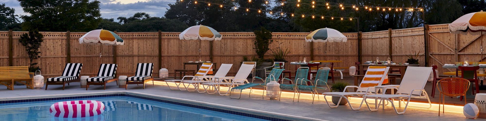 A night-time pool scene with string lights, a row of lounge chairs, umbrellas, and a calm blue pool under a partly cloudy sky.