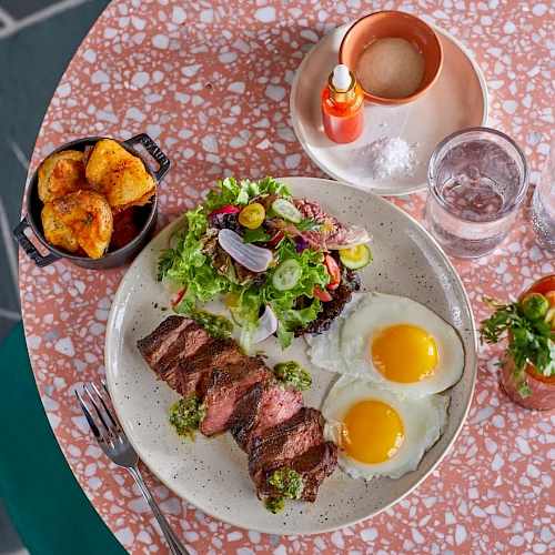 Two sunny-side eggs with steak and greens on a plate, plus a side of crispy potatoes and a small salad, on a pink terrazzo table with a candle.
