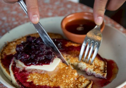 A plate of pancakes topped with jam and butter, being cut with a knife and fork, on a patterned table. End.