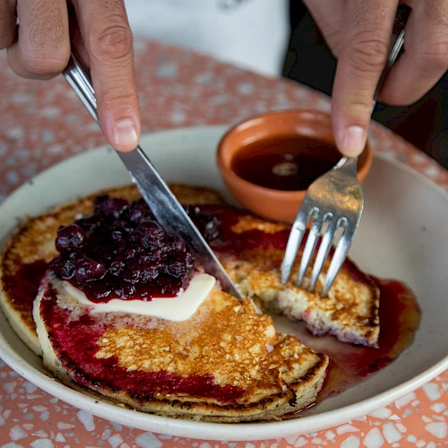 A plate of pancakes topped with jam and butter, being cut with a knife and fork, on a patterned table. End.