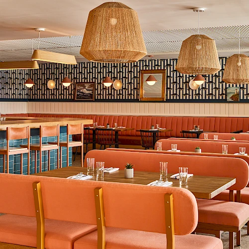A retro-style diner with peach-pink booths, wooden chairs, and woven hanging lamps; a long bar, tiled wall, and cozy retro ambiance.