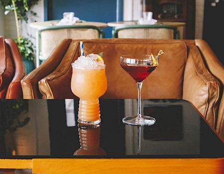 Two drinks, one orange and one dark red, sit on a black table in front of leather chairs in a relaxed setting with plants in the background.