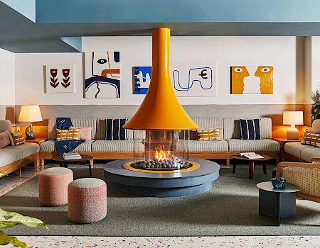 A modern living room with a central fireplace, surrounded by sofas and colorful cushions, art pieces on the walls, and soft lighting.