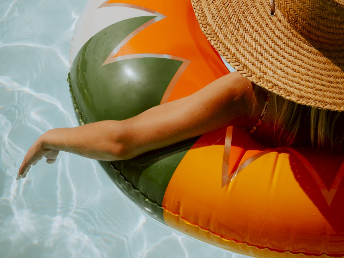 A person floats in a pool on a colorful inflatable ring with a sunburst pattern, wearing a wide straw hat.
