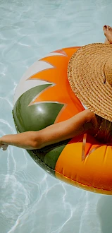 A person wearing a straw hat relaxing on a colorful inflatable ring in a clear pool, with one arm resting on the edge.