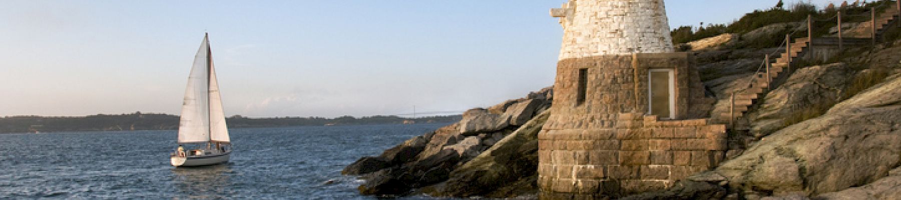 A lighthouse stands on a rocky shore, overlooking a sailboat on the ocean, under a clear sky with gentle waves hitting the rocks.